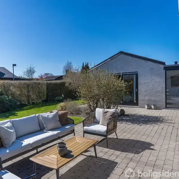 Terrassemøbler står på en flisebelagt terrasse. Sofa og stole omkring et kaffebord. I baggrunden ses et gråt hus, en grill og grønne buske under en klar blå himmel.