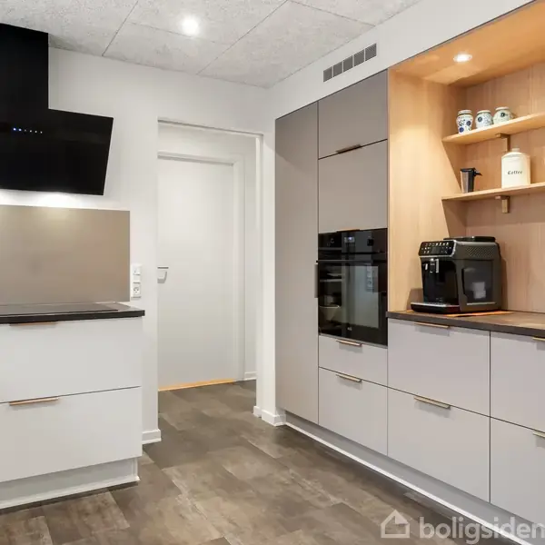 Køkken med moderne skabsløsning, indbygget komfur og åbne hylder. Hylder med dåser og glas. Til højre ses en kaffemaskine på en bordplade. Lysegråt gulv og hvidt loft. Ingen tekst fundet.