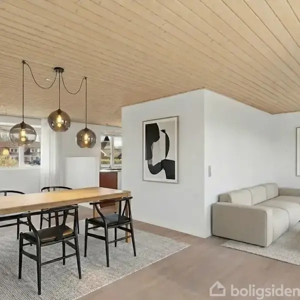 Et moderne stue/køkken-alrum med træloft. Et spisebord med fire stole står under tre hængende lamper. En sofa med to abstrakte billeder på væggen i baggrunden. En plante til højre.