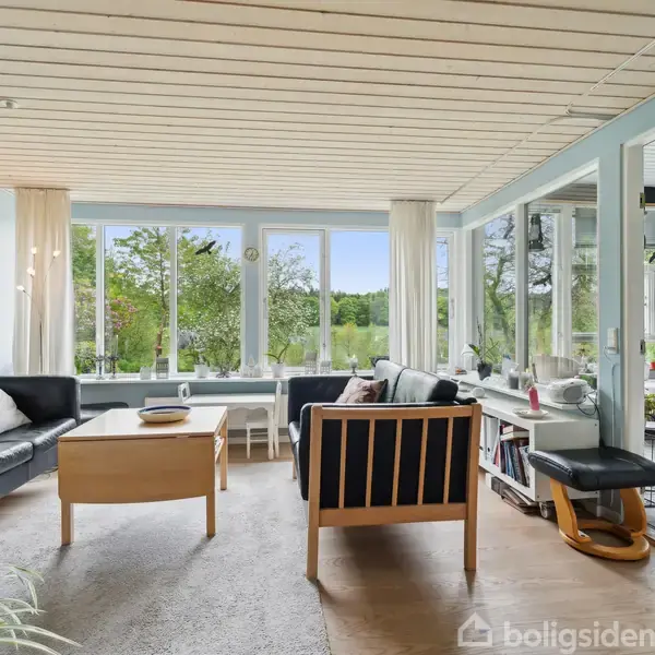 En stue indeholder sorte lædersofaer og et træsofabord, beliggende i et lyst rum med store vinduer. Udsigt ud over en grøn have skaber en rolig atmosfære.