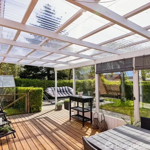 En overdækket terrasse med trægulv indeholder møbler og planter. I baggrunden ses en have med grønne buske og et drivhus, mens solen skinner gennem et gennemsigtigt tag.