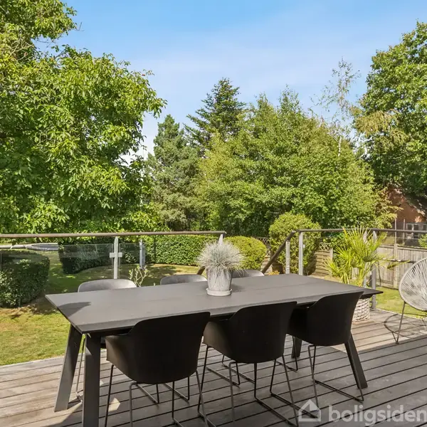 Et stort udendørs spisebord med sorte stole står på en terrasse med glasrækværk. Det er omgivet af frodige grønne træer og buske i en have.