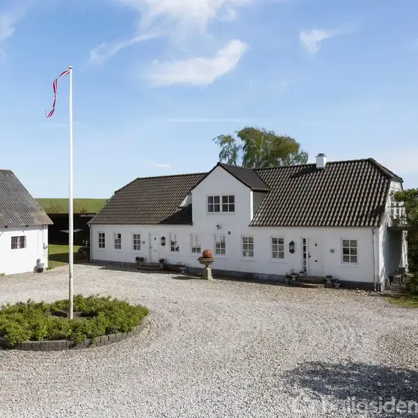 Hvidt hus med gråtag står ved siden af en lys gårdsplads med en flagstang og grusbelægning, omgivet af grønne træer og en klar blå himmel.