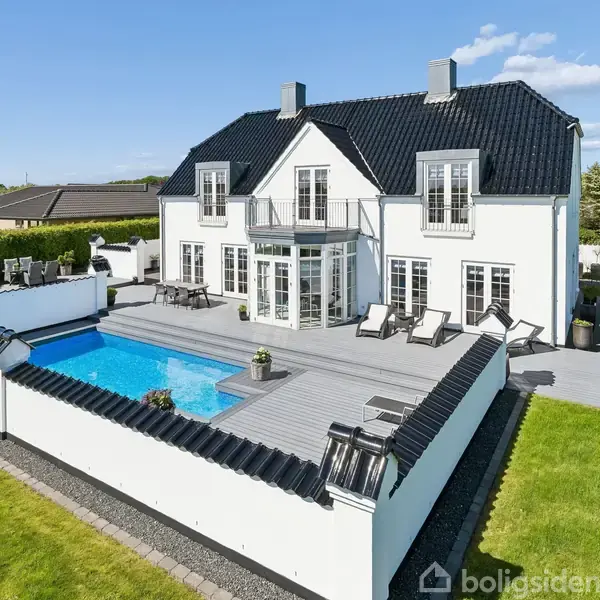 Hvid to-etagers hus med sort tag står foran en terrasse med liggestole. En rektangulær swimmingpool er omgivet af lave hvide mure. Græsplæne omgiver området under en blå himmel.