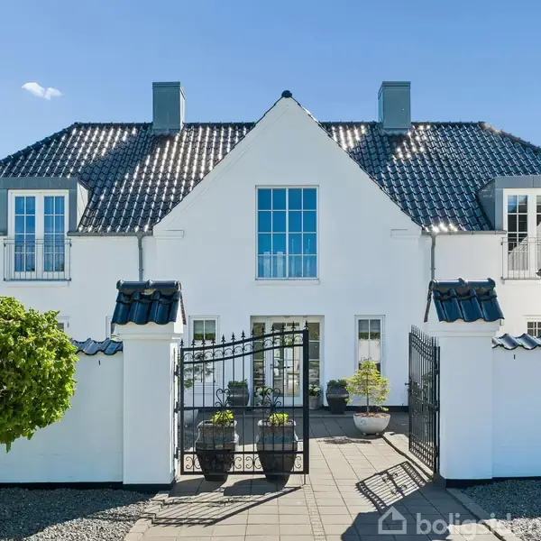 Hvidt hus med sort tag og store vinduer står majestætisk. Jernporten åbner ind til en flisebelagt gård. Et træ står til venstre, omgivet af småsten.