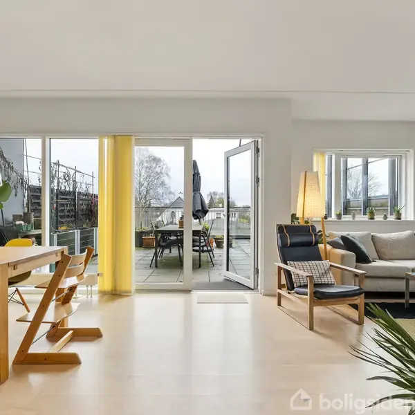 En lys stue med spisebord og stole til venstre, sofa og stol til højre, der åbner ud mod en altan med borde og stole. Planter tilføjer grønne detaljer.