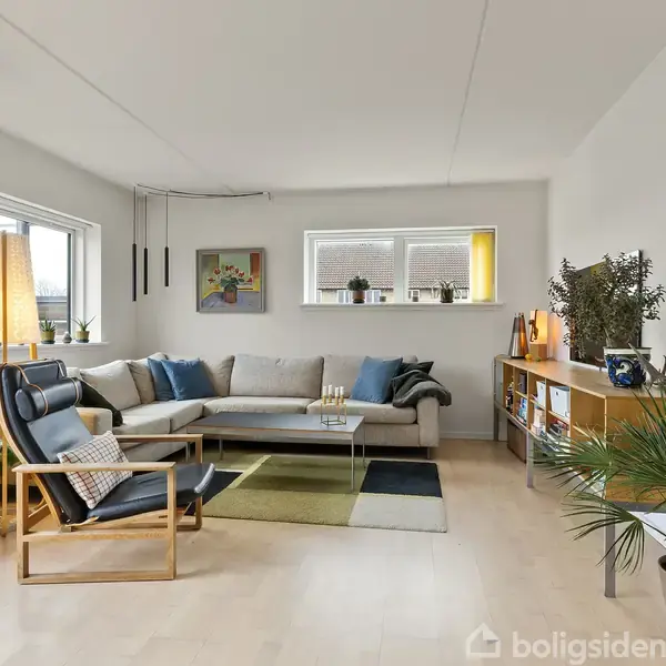 En hjørnesofa er placeret i et lyst, moderne rum med træmøbler og planter. Et sofabord står foran sofaen, mens kunst og hylder pynter væggene. Gulvlampe til venstre.