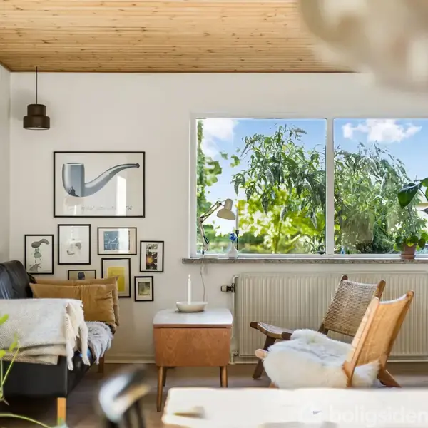 Sofa med tæpper står ved væg dekoreret med billeder, mens lænestol og bord med lys er placeret foran stort vindue med udsigt til træer og blå himmel.