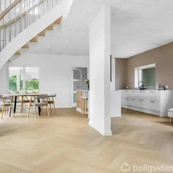 En moderne stue med en hvid, svungen trappe til venstre. Et spisebord med stole står centralt, mens et køkkenområde med hvide skabe og en sofa er til højre.