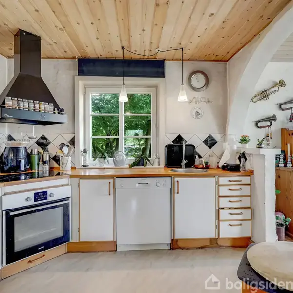 Køkken med hvide skabe og træbordplader. En ovn med kogeplader står til venstre, og en emhætte er monteret på væggen. Vindue centralt med udsigt til træer.