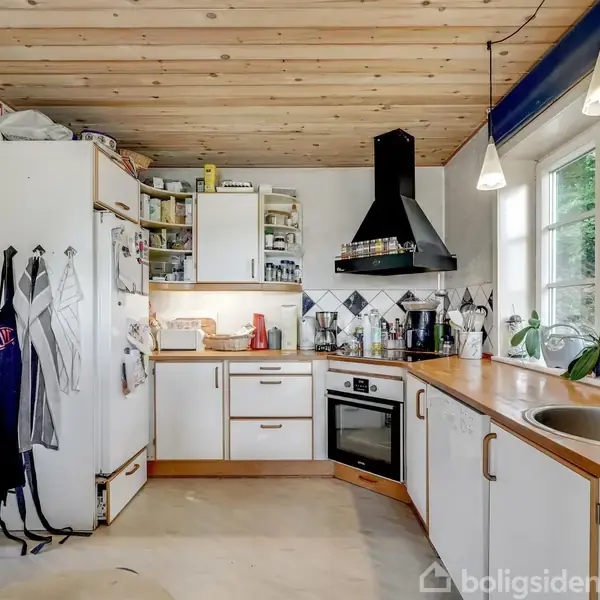 Et køkken med hvide skabe og træbordplade har opvask, krydderier og en brødrister ved vinduet. En emhætte hænger over komfuret. Kontekst: Træloft og lysekrone.