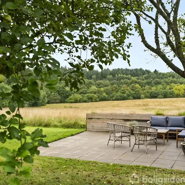 Terrasse med havemøbler står på flisebelagt område. Møblerne består af sofa, stole og borde. Omgivet af grønne marker og træer, hvilket skaber en fredfyldt, naturlig atmosfære.