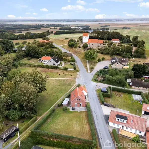 Et fugleperspektiv viser landlige omgivelser med spredte huse og en kirke i centrum omgivet af marker og træer. Veje forbinder bygningerne i det åbne landskab.