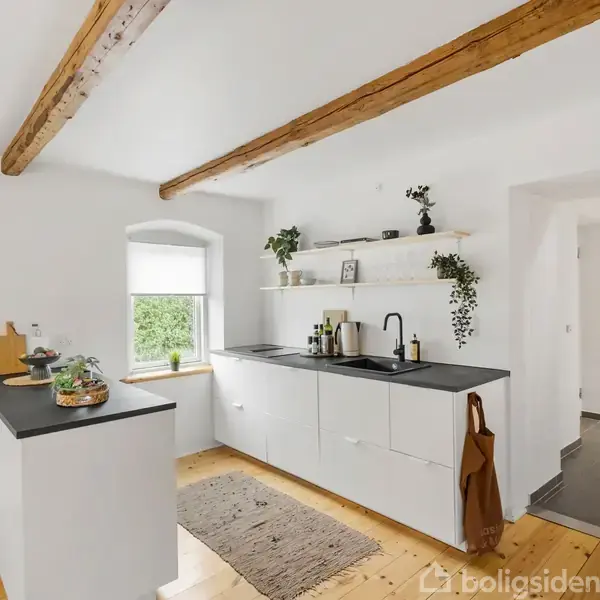 Et lyst, moderne køkken med hvide skabe og en sort bordplade. Hylder med planter og dekoration hænger over vasken. Træbjælker i loftet tilføjer en rustik stemning.
