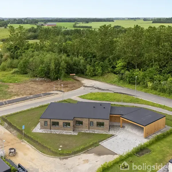 Et moderne hus med fladt tag står stille i en forstadsområde omgivet af grønne marker og skove. En brolagt indkørsel leder til en carport ved siden af huset.