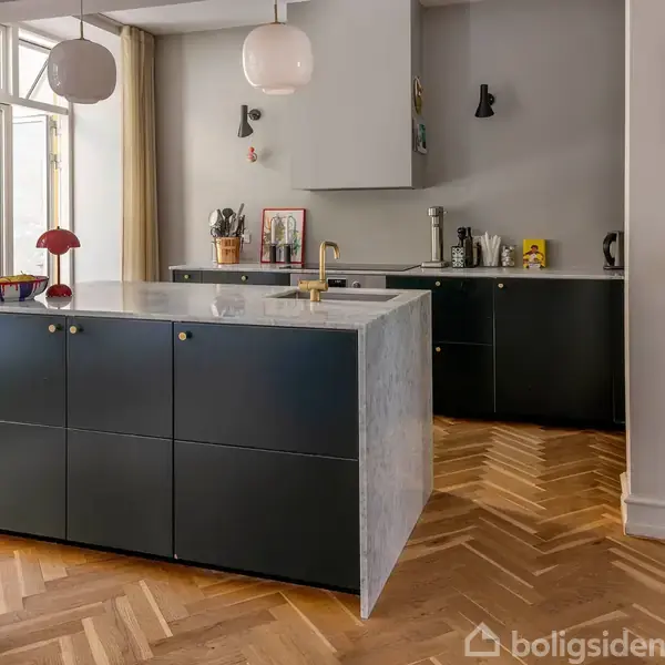 Køkkenø med sort kabinet og marmorbord fungerer som rumdeler foran beige gardiner. Omgivet af træparketgulv, vægkunst, og hængelamper. Baggrund har ekstra skabsplads og køkkenudstyr.