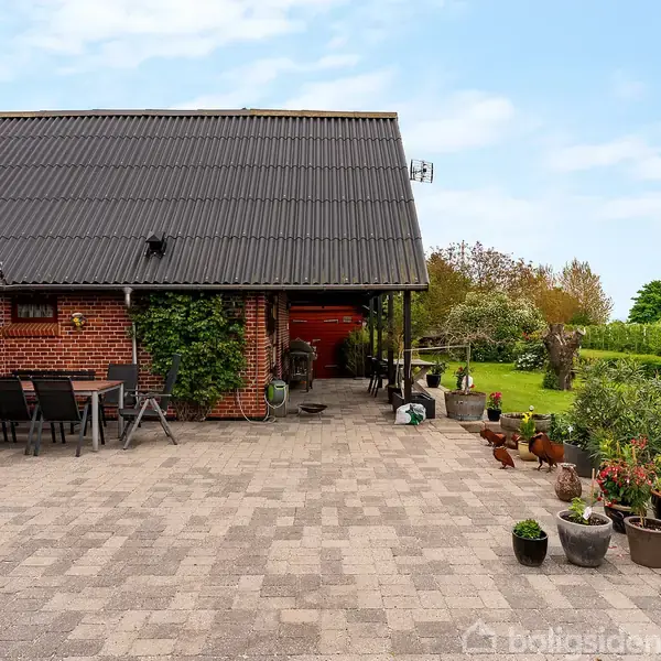 Rødt murstenshus med sort tag står ved siden af en stor flisebelagt terrasse. Bord og stole står til venstre. Planter i krukker kjeder terrassen. Græsplæne og buske til højre.