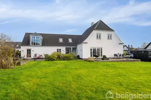 Et hvidt to-etagers hus står stille på en stor, velplejet græsplæne. Ved siden af står en cykel, og terrassen er møbleret til udendørs brug.