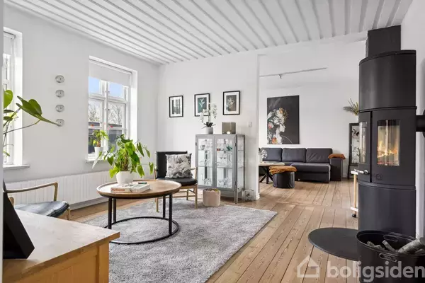 En lys stue med lyse trægulve, en rund sofabord omgivet af en sofa og en pejs i hjørnet. Grønne planter og moderne kunst dekorerer rummet.