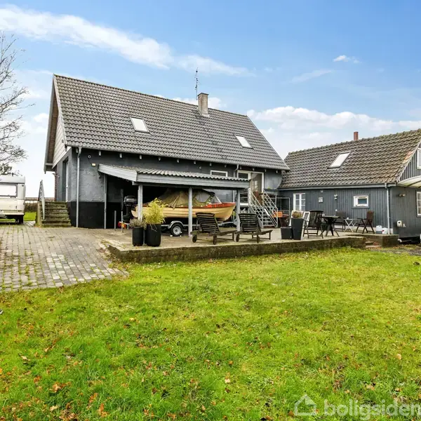 En stor, grå bolig med skråtag står i en grøn have med en parkeret hvid bil og trailer. Terrassen har stole og borde. Skyfri blå himmel i baggrunden.