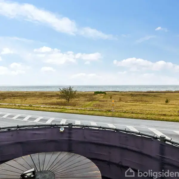 Terrasse med rundt bord foran en kystudsigt. Vejen strækker sig langs kysten med fladt, gult græsland og enkelte træer. Himlen er blå med få skyer.