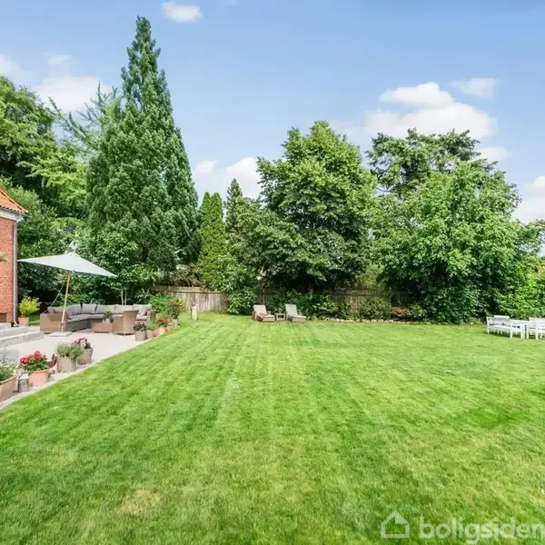 En rød murstensvilla med terrasse og trapper fører ned til en stor græsplæne omgivet af frodige træer. Havemøbler står ved siden af huset.