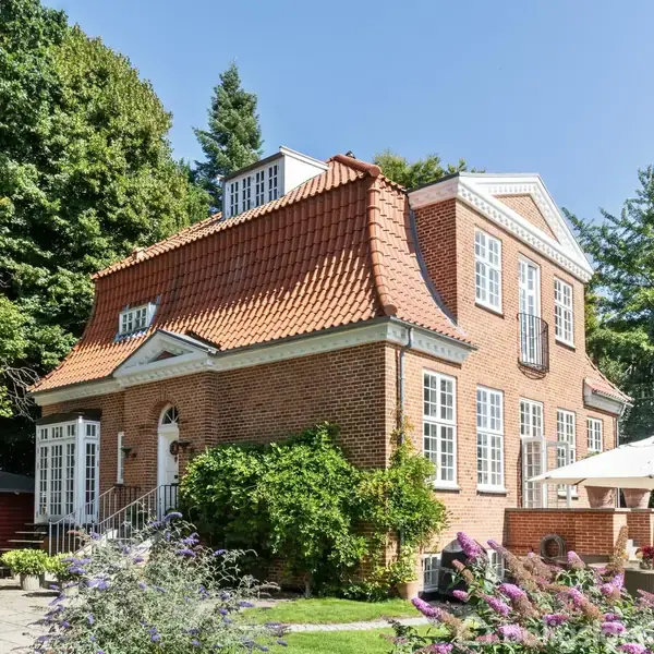 Rødt murstenshus med klassisk tegltag står stille ved en terrasse med parasoller, omgivet af frodig have med høje træer og blomsterbede i et solrigt, roligt område.