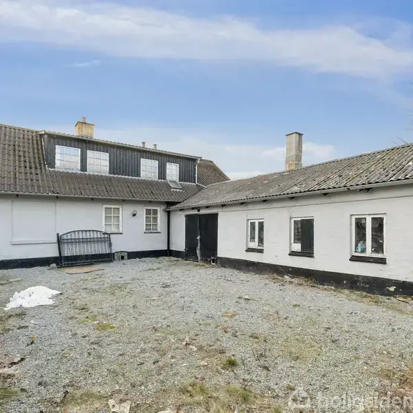 Hus med hvid facade og sort tag står i en gårdsplads omgivet af sten og et sort hegn, enkelt træ til højre, klar himmel i baggrunden.