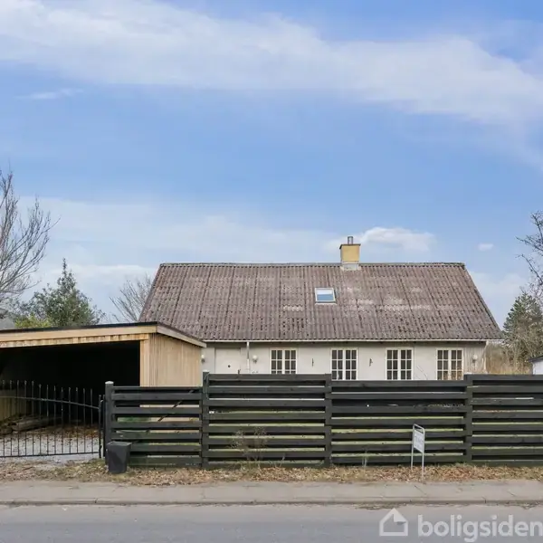Et lille hus med rødt tag står bag et sort plankeværk og en carport. Træer omgiver ejendommen under en blå himmel.
