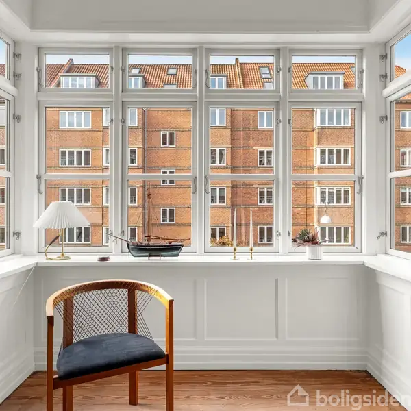 En stol står ved et stort vindue med udsigt til en murstensbygning. En lampe og en modelbåd er placeret på vindueskarmen i det lyse rum.