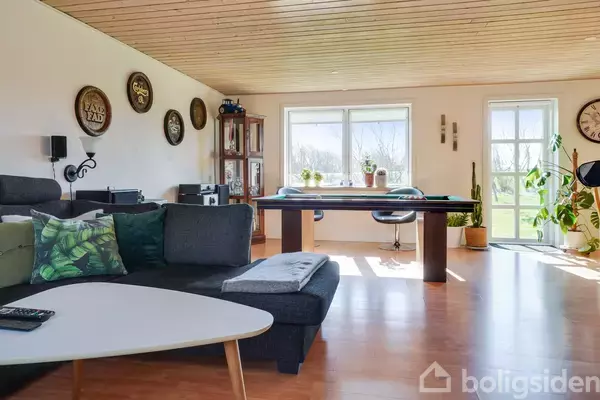 En moderne stue med sort sofa og grønne puder, et spisebord ved vinduet, dekorative vægplader, planter og en rejsekuffert i hjørnet, mens sollys strømmer ind.