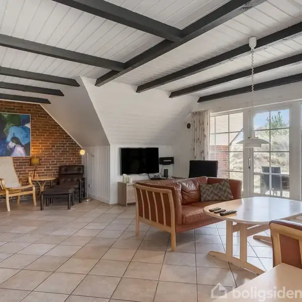 Stue med sofa og borde, set fra et hjørne. Store vinduer lukker lys ind. Der er synlige loftsbjælker og teglstensvæg. Kunst hænger på væg. Pejs ved stolene.