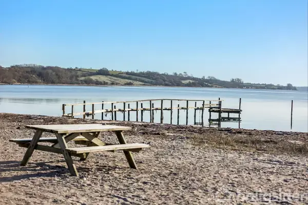 En træbænk står stille på en sandstrand med udsigt over en rolig sø. En træbro strækker sig ud i vandet, omgivet af bakkede landskaber under en klar blå himmel.