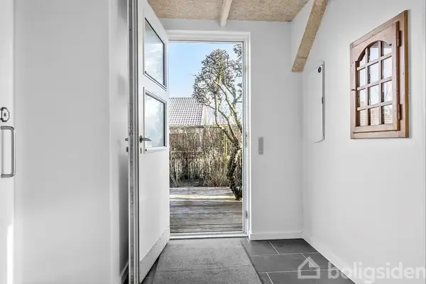 En hoveddør står åben og leder ud til en terrasse med udendørs træ og buske. Indenfor er der lyse vægge, et lille vindue og gulvfliser.