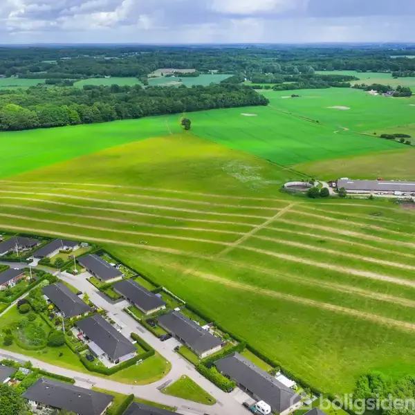 Landsbyhuse grænser op til en stor, stribet mark i et landområde. Rundt om ses åbne marker og skove under en overskyet himmel.