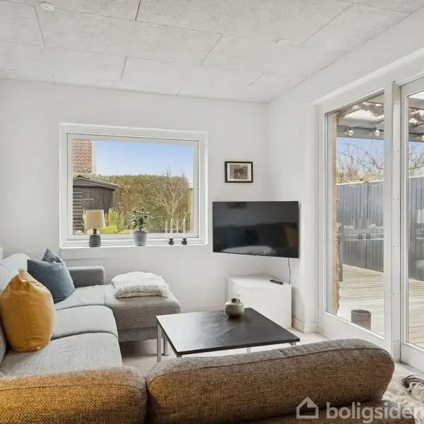 En stue med en grå hjørnesofa og et sofabord placeret foran et fladskærms-TV. Vinduer og terrassedør giver udsigt til en udendørs terrasse med grill og træhegn.