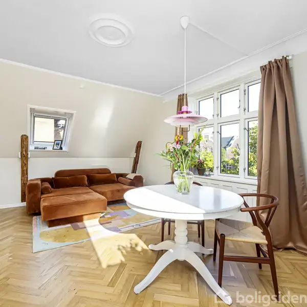 En lys stue indeholder en rund, hvid spisebord med en vase med blomster stående på det. En brun sofa står nær et vindue med trærammer og brune gardiner.