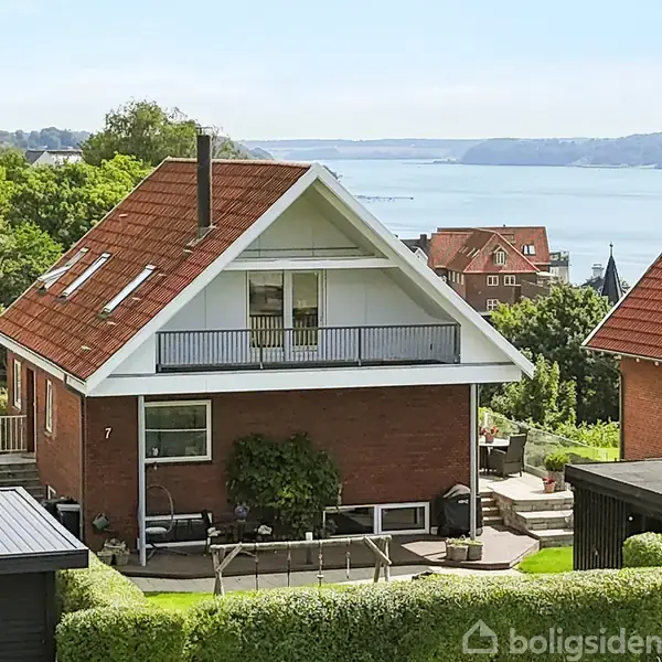 Et to-etagers murstenshus med rødt tag ligger omgivet af grønne hække. Huset har en stor veranda og udsigt over en fjord med skovklædte bakker i baggrunden.