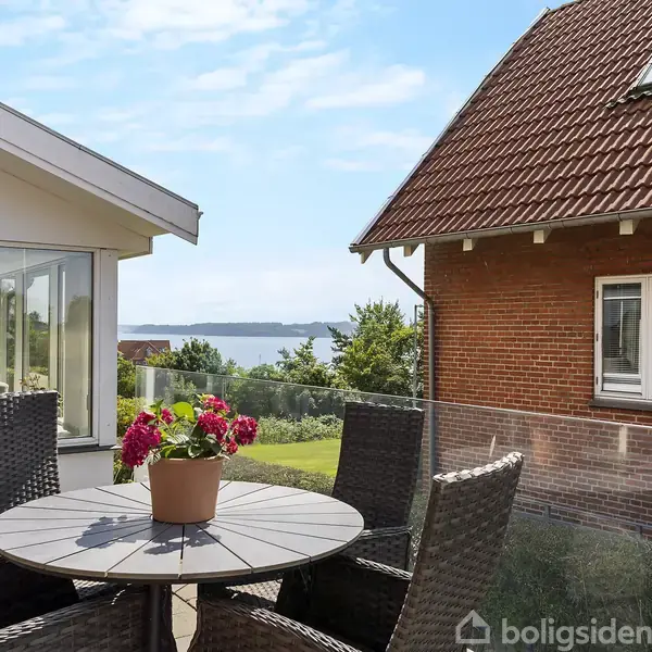 Et rundt bord med en potteplante står på en terrasse med udsigt over vandet, omgivet af fletstole. En moderne bygning og en murstensvilla ses i baggrunden.