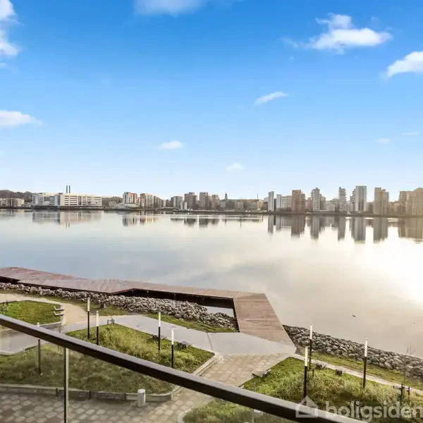 Udsigt over stille vand fra en balkon eller terrasse, med moderne bygningskompleks i baggrunden. Himlen er klar med få skyer, og området er omgivet af græs og promenade.