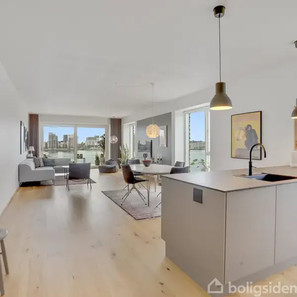 Bygning består af moderne lejligheder med store glasfacader. To parallelle strukturer i centrum flankeres af højere bygninger med altaner. Omgivet af flisebelagt gård og grønne planter.