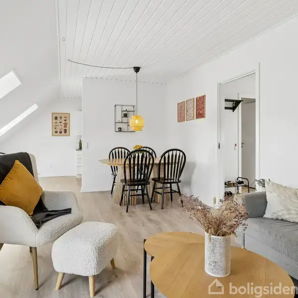 En lys stue med en grå sofa og et rundt sofabord. En lænestol med gul pude står ved siden af. I baggrunden ses en spisestue med sorte stole.