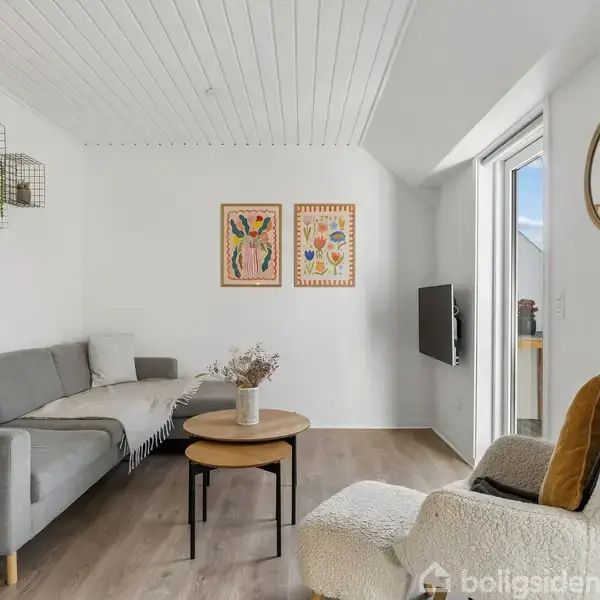 Stue med en grå sofa i hjørne, ved siden af et rundt sofabord med vase. Kunst hænger på væggen. En lænestol står ved en dør til altanen. Skylight skaber lys.