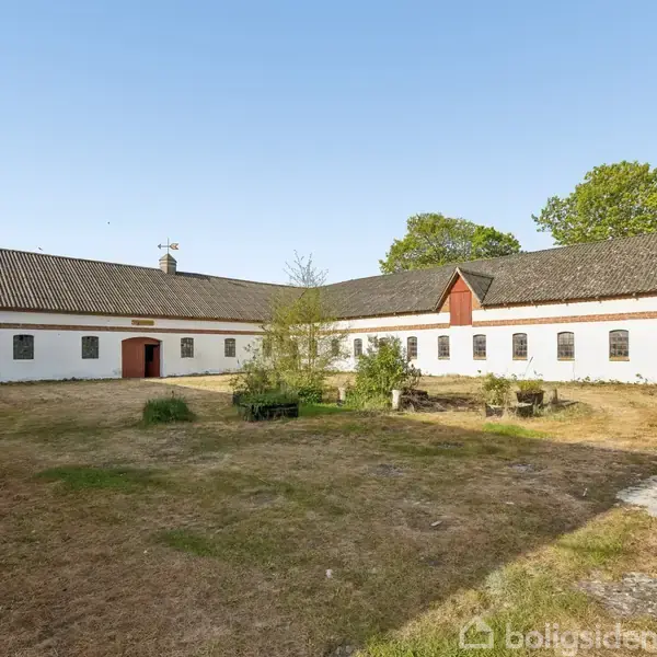 En hvid landbygning med rødt tagstensloft står stille i en gårdsplads med græs og buske, omgivet af træer under en klar blå himmel.