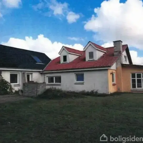 En hvid og gulmalet villa med rødt tag står på en græsplæne under en blå himmel med få skyer. Flere vinduer og en skorsten er synlige.