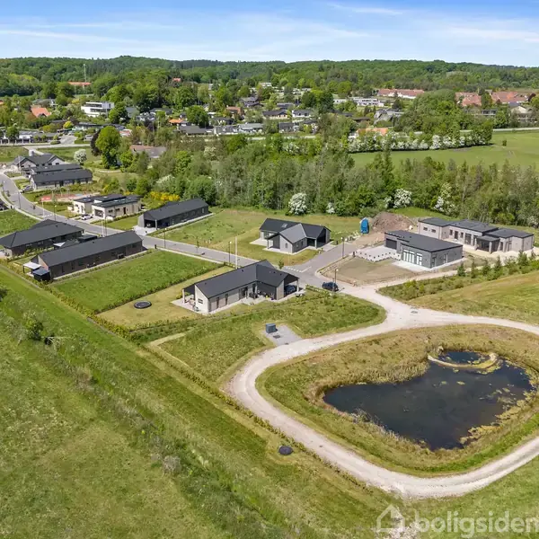 Boligområde med moderne huse omgivet af grønne marker. En lille sø er placeret midt i området. Omkringliggende landskab består af træer og en fjernere by i baggrunden.