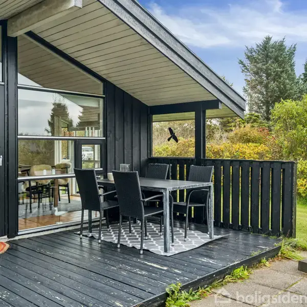 Sortmalet træt terrassebord med fire stole står på en sorte træveranda. Glasdøre åbner til indendørs spisestue. Omgivet af frodig grøn have og buske under en blå himmel.