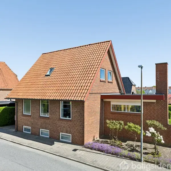 Et rødt murstenshus med rødt tegltag står på en villavej. Huset har flere vinduer, en skorsten og en lille have med blomster ved vejen.