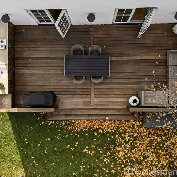 Træterrasse med spisebord og stole i midten, omgivet af sofa og udendørs køkken. Efterårsblade ligger spredt på græsplænen omkring terrassen.