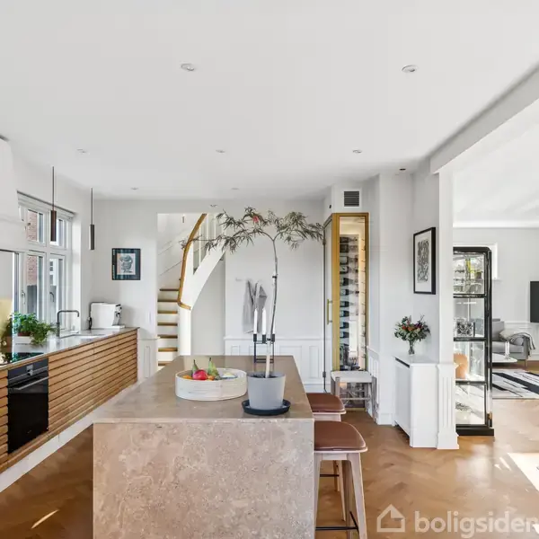 Et moderne køkken-alrum med en køkkenø af marmor, brune trælåger og hvide vægge. I baggrunden er en trappe, og stuen ses med en sofa og TV.